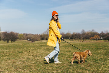 a girl in a yellow jacket and a red hat with glasses walks with a spaniel dog in the park and speaks on the phone