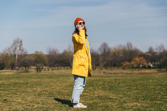 a girl in a yellow jacket and a red hat with glasses stands sideways in full growth in the park and looks at the smartphone screen