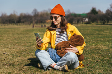 Naklejka na ściany i meble a mother in a yellow jacket and a red hat with glasses sits on the grass in the park and looks at the screen of the phone in her hands, holds a baby in her hand and breastfeeds, a spaniel dog sits nea
