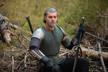 A gray-haired male warrior with a beard and a scar. In armor and with a sword, he sits on a log. An old knight at a halt with a weapon in his hands.
