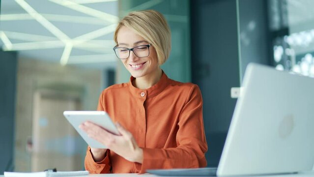Happy young blonde woman using tablet while sitting at workplace at desk in modern office. Smiling female employee wearing glasses browses social networks, reads email messages, chats online