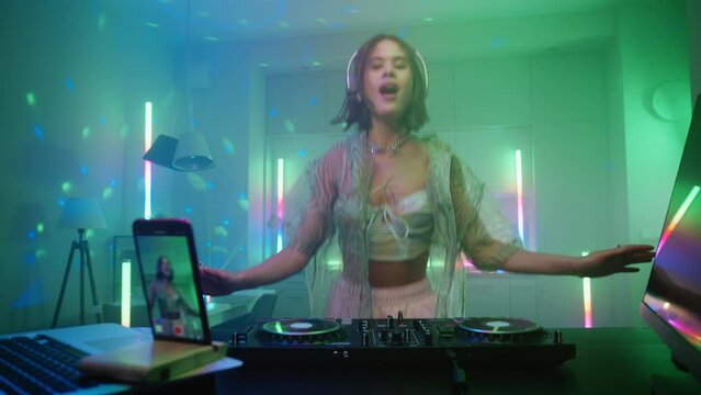 Beautiful young stylish woman dancing enjoying her live dj set in home interiors at night, excited female in headphones recording electonic music on turntable console. Colored neon lights glow