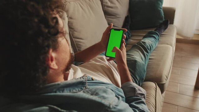 Young man lying on sofa uses mobile phone with green mock-up screen chroma key