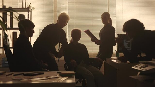 Silhouettes of a young management team are discussing a new project or report. A youth group of men and women work together in a dark office in the evening near a backlit window. Teamwork.