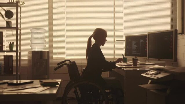 Silhouette of a young woman in a wheelchair typing on a computer keyboard and drinking coffee in a dark office in the evening. A disabled female manager works sitting at a table near a backlit window.