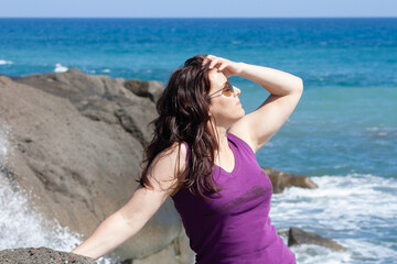 woman with sunglasses posing on the beach and leaning on rocks in the background you can see the sea