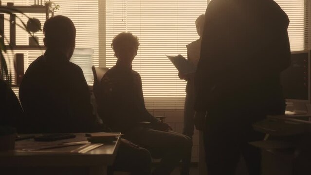 Silhouettes of a young management team are discussing a new project or report. A youth group of men and women work together in a dark office in the evening near a backlit window. Teamwork.