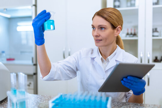 Female lab scientist working on samples and make notes on clipboard, holding flask with blue liquid