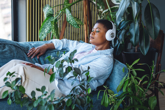 Young beautiful African American woman relaxing on the sofa at home among green plants listening to music in headphones. Meditation, relaxation, mental health concept. Enjoying life, happiness, calm