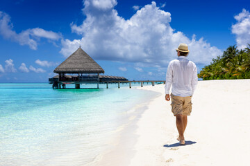 A man in white clothing and sunhat walks down a tropical beach in the Maldives islands and enjoys...