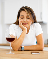 Upset young woman with glass of red wine in kitchen reading messages on mobile phone screen