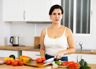 Obraz na płótnie Canvas Smiling young Hispanic woman standing in home kitchen, preparing fresh vegetable salad. Vegetarian diet concept