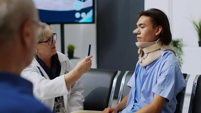 Asian patient with neck collar doing consultation with elderly medic, doing concussion exam to check vision and brain damage with light. Person attending checkup appointment with medic.