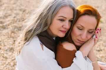 An elderly gray-haired mother and her adult daughter sit on the shore and cuddle. Caring for an elderly mother and the concept of a happy retirement and old age. close up