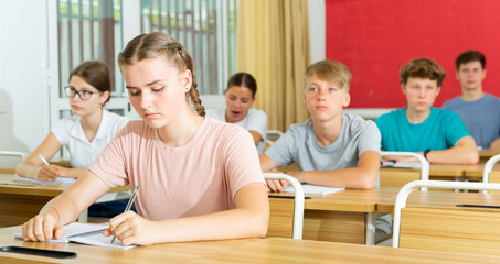 Portrait of focused teenage schoolgirl writing lectures in workbooks in classroom during lesson..