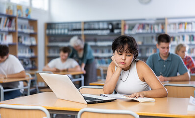 Portrait of tired or bored teenage female pupil sitting at desk in library during lesson
