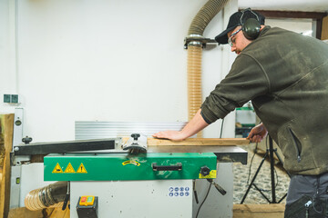 Focused male carpenter in protective headphones and eyewear using wood surface planer machine....