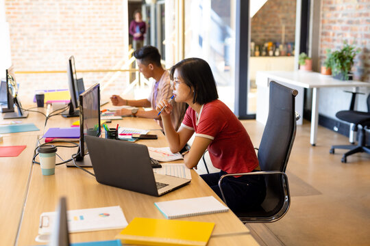 Thoughtful asian female creative working on computer at desk in creative office with male colleague