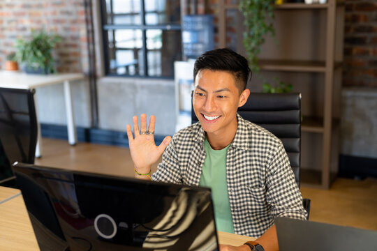 Smiling asian male creative at computer making video call, waving in casual office