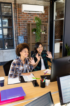Happy diverse female creatives at computer, making video call, waving in casual office, copy space