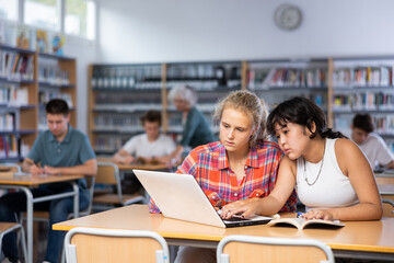 Fototapeta na wymiar Portrait of asian schoolgirl with her spanish female friend studying in library using laptop