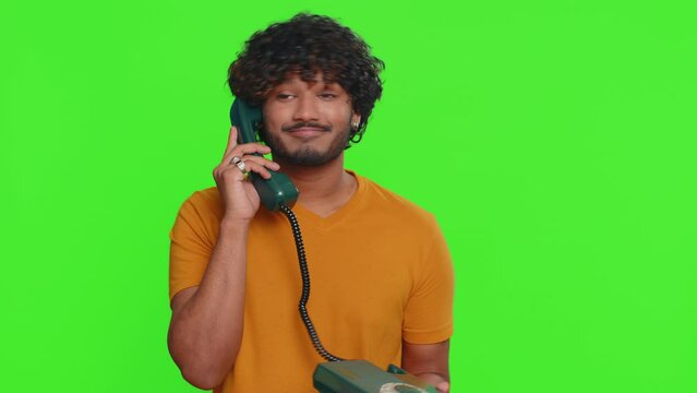 Crazy funny young indian man talking on wired vintage telephone of 80s, fooling, making silly humorous faces. Communication, conversation talk. Hindu hacker guy isolated on green chroma key background