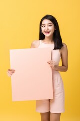 Fototapeta na wymiar Cheerful Asian woman dressed in pale pink clothes holding up an empty signboard. Her friendly smile, against a yellow background is perfect as content mockup. Created with generative A.I. technology.