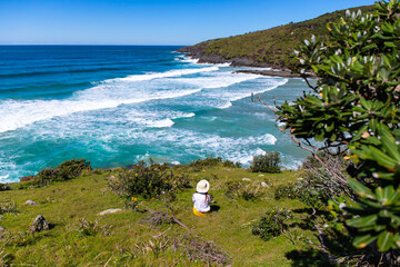 Girl in a hat sits and looks at stunning hidden third beach (cove) in Hat Head National Park,...
