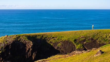 girl in white shirt and walks along the grassy cliff gazing at the sea in hat head national park in...