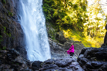 hiker girl sitting on rocks in front of large tropical waterfall in australian rainforest; red...