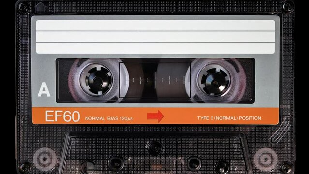 Audio cassette tape in use for sound recording in the tape recorder. A vintage, orange, brand new blank labelled music cassette playing back in a deck player. Static video camera shot. Close up, 4K
