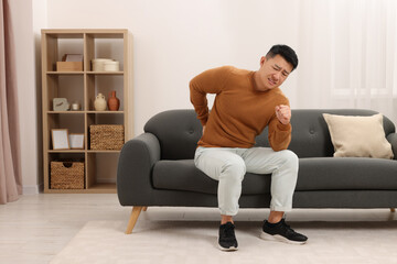 Asian man suffering from back pain on sofa indoors