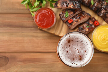 Glass of beer, tasty grilled ribs and sauces on wooden table, flat lay. Space for text