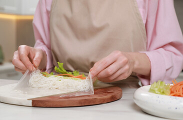 Making delicious spring rolls. Woman wrapping ingredients into rice paper at white table, closeup