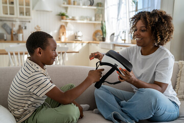 African American mother assisting son with virtual reality glasses at home, helping to use headset safely, happy mom and kid sit together on sofa exploring virtual world. Child and vr experience