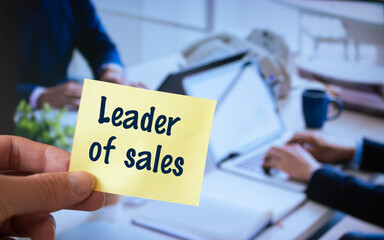 Male hand holding sticky note written Leader of sales.