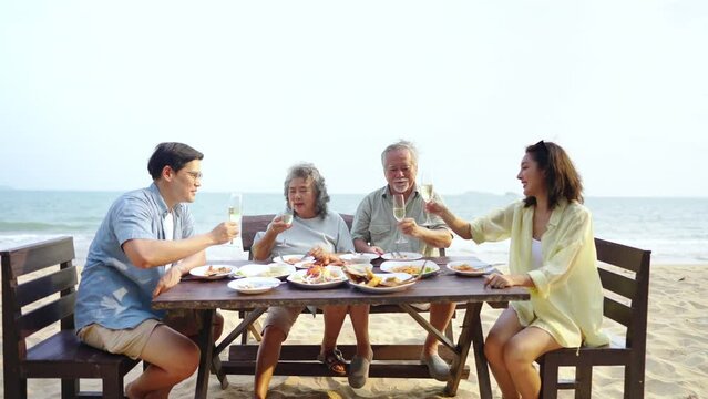 Happy Asian couple and aging parents having celebration dinner and drinking champagne together at tropical beach restaurant during travel ocean on summer holiday vacation. Family relationship concept.