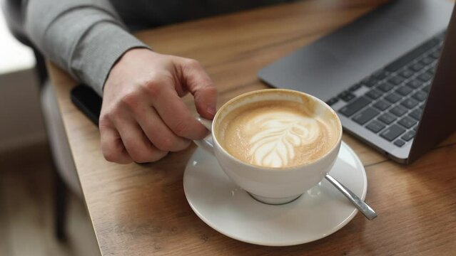 Video of sporty businessman drinking cream coffee stirring with teaspoon near expensive grey macbook on table in office