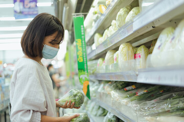 Portrait of healthy young Asian woman with casual clothes is wearing medical face mask when buy fresh organic vegetable to make healthy food with new normal lifestyle, social distancing at supermarket