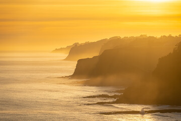 Aerial view of some cliffs in the sea in a sunset of orange colors and mist over the water, Asturias, Spain.