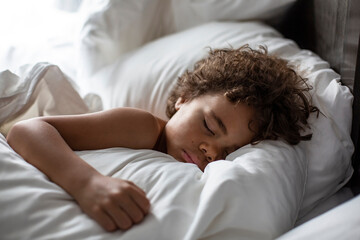 Cute African American little boy sleeping soundly in his nice warm bed with white sheets and pillowcase in the morning. 