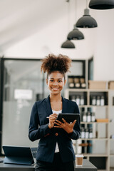 Fototapeta premium Confident beautiful African businesswoman typing laptop computer and digital tablet while holding coffee at office.