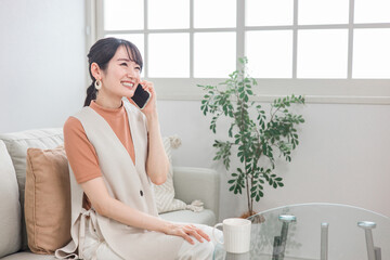 ソファに座ってスマホで友達と電話する笑顔のアジア人女性
