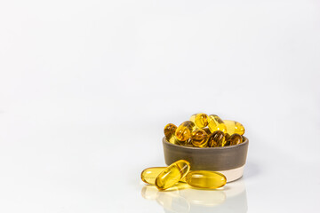 A close-up photo of several yellow capsules of fish oil in a small bowl on a white background. Copy space, reflection.