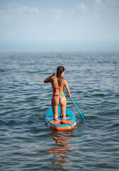 Attractive slim tanned Caucasian brunette woman in a bikini with her back to the camera paddling on her knees on her paddle surf board in the water of the sea.
