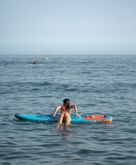 General shot of an attractive, slim brunette woman with her back to the camera trying to get on her surfboard in the sea water.