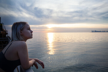 Obraz premium Beautiful blonde woman in tank top standing on pier on the background of city silhouette, cloudy sky and sea