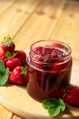 Strawberry jam in glass jar on wooden board with fresh strawberry fruit and green leaves on wooden background. Recipe of delicious homemade berry jam of strawberry full of vitamins and antioxidants.