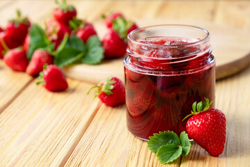 Strawberry confiture in glass jar on wooden board with fresh strawberry fruit and green leaves on wooden background. Recipe of homemade berry jam of strawberry full of vitamins and antioxidants.