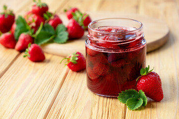 Strawberry jam in glass jar on wooden board with fresh strawberry fruit and green leaves on wooden background. Recipe of delicious homemade berry jam of strawberry full of vitamins and antioxidants.